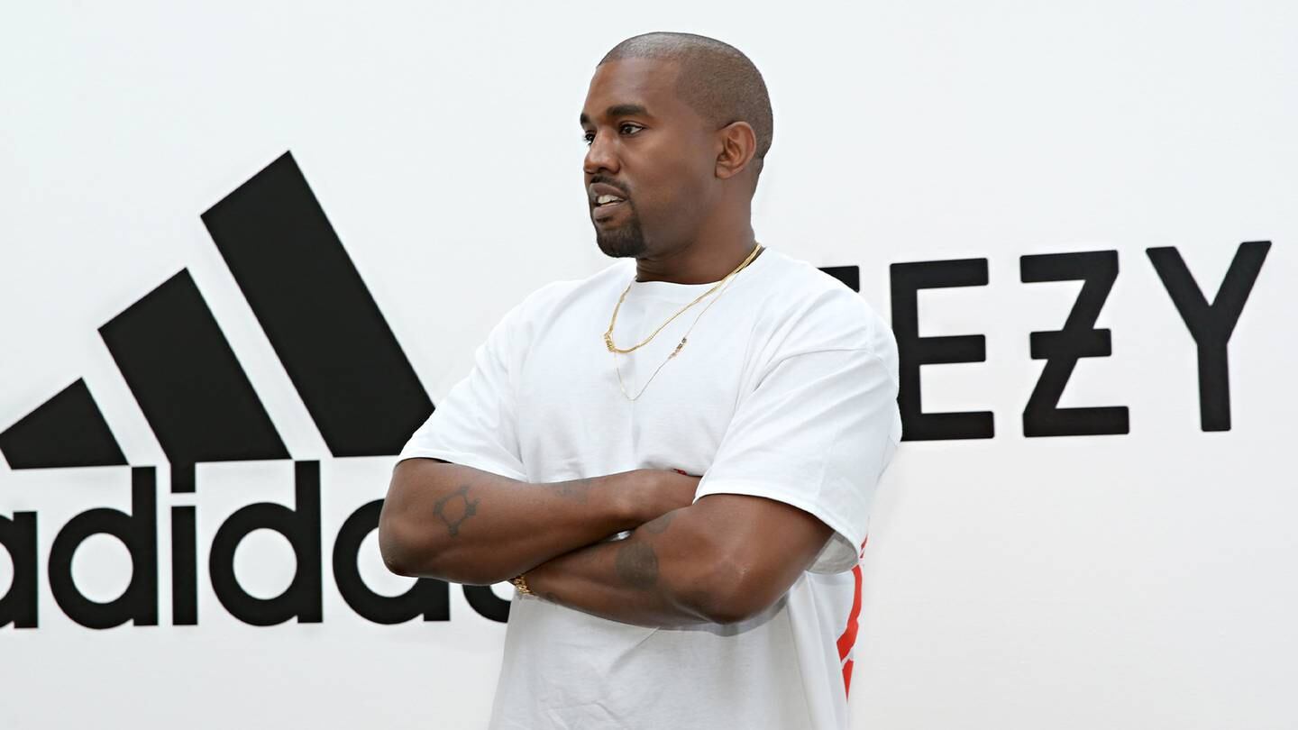 Rapper Ye standing in front of an Adidas and Yeezy photo wall with the logos behind him.