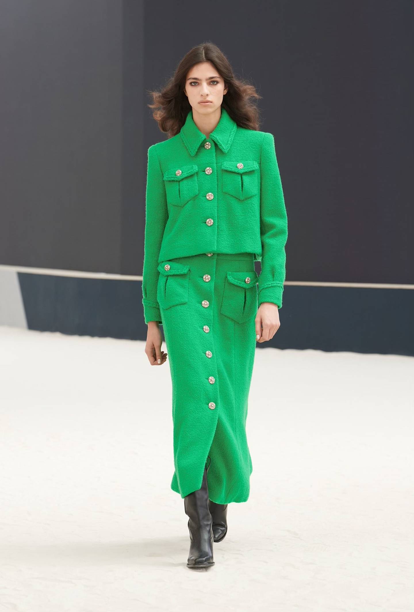 A model wears a green long sleeved blazer with a matching maxi skirt with gold buttons down the middle. The tweed outfit is paired with black heeled boots.