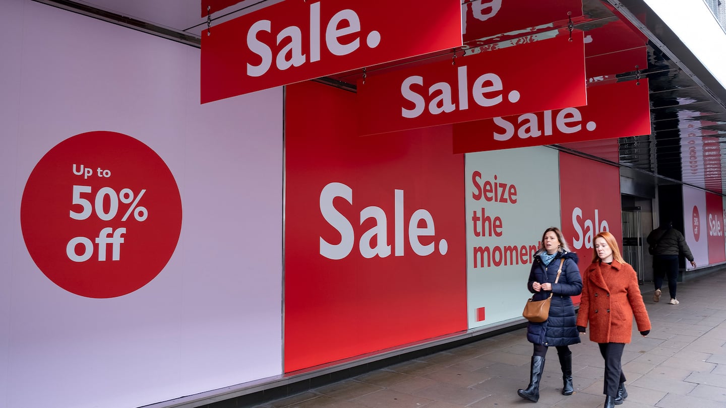 People out shopping on Oxford Street walk past large scale January sale signs in red and white for major high street clothing retail shops.