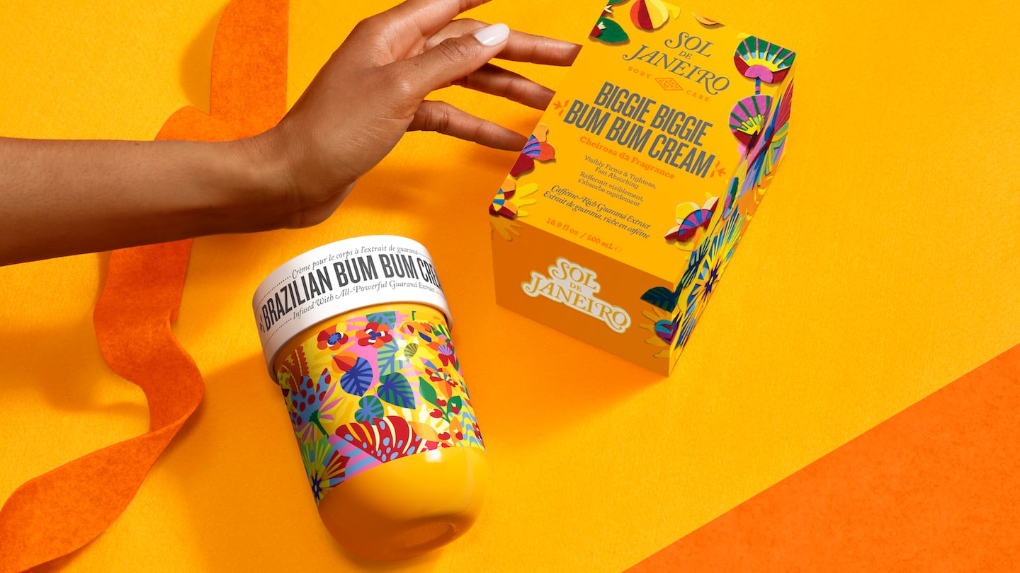 A container of Sol de Janeiro's Brazilian Bum Bum Cream with a person's hand holding a box.