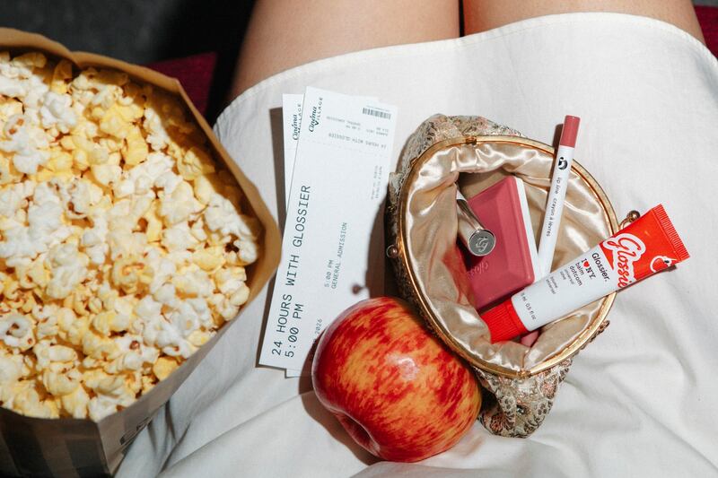 A Glossier makeup bag, apple and popcorn photographed on the lap of somebody watching a movie at a theatre