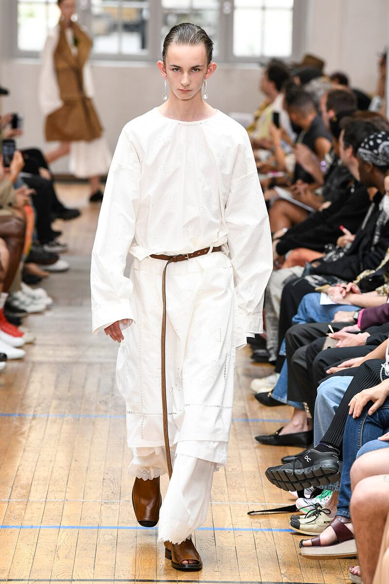 A model wears a relaxed fit white outfit with a long shirt and brown long belt and shoes.