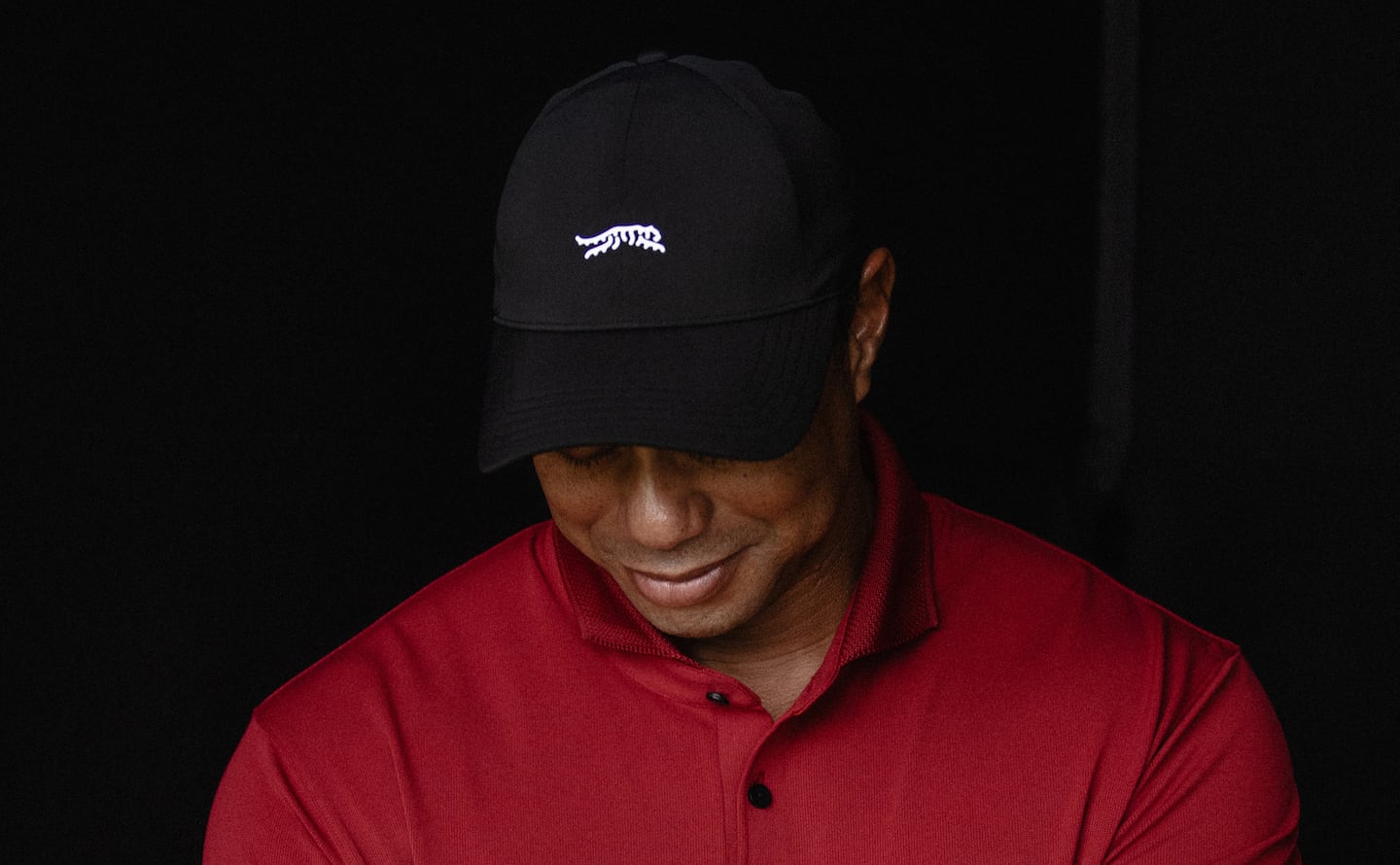 Tiger Woods in a black hat with the tiger logo of his new brand in white, wearing a red polo shirt from the brand.