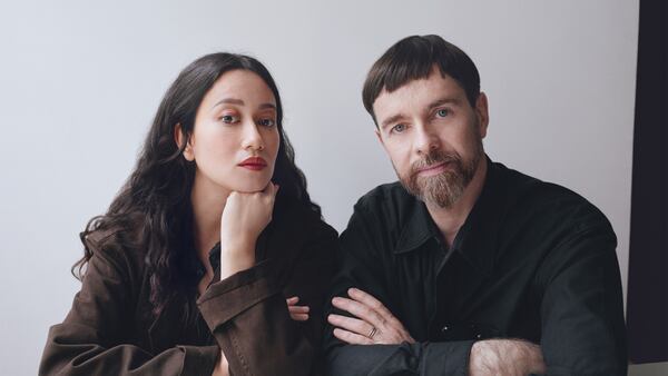 A portrait of two people. The woman on the left wears a brown jacket and rests her head on her hand. The man on the left has his arms folded.
