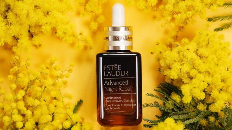 Estée Lauder advanced night repair serum bottle laying on a yellow background with yellow flowers surround it.