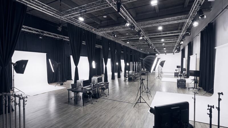 A large-scale commercial photography studio featuring a row of modular shooting bays equipped with white seamless backdrops and professional lighting. Each station is paired with a tethered iMac workstation on a polished wood floor, flanked by black floor-to-ceiling privacy curtains and an overhead industrial lighting truss.