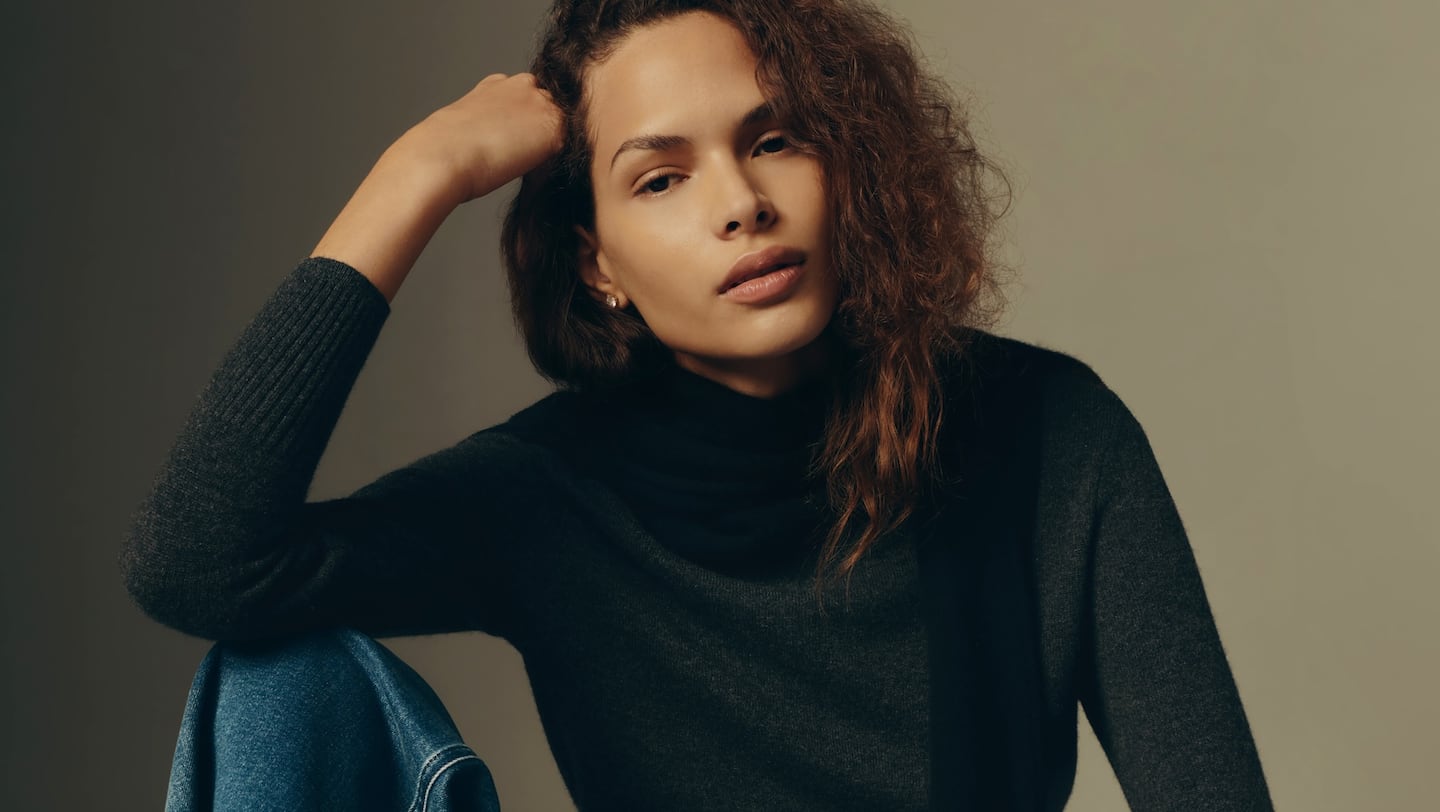 A woman sits with her elbow on her knee in a grey high-neck sweater and jeans by Quince, which is launching in the UK.