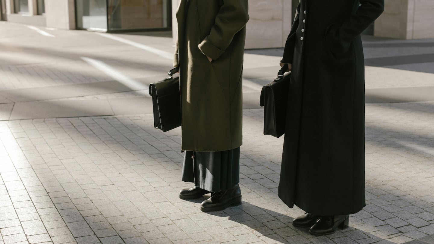 An image shows two people standing side-by-side on a cobblestone plaza in direct sunlight. Both are shown from the neck down, wearing long, structured overcoats and carrying black leather briefcases. The person on the left wears an olive green wool coat over a long, pleated dark skirt and chunky black boots. The person on the right wears a floor-length black coat with white buttons and black leather heeled boots. Long shadows stretch across the light-colored pavement behind them.