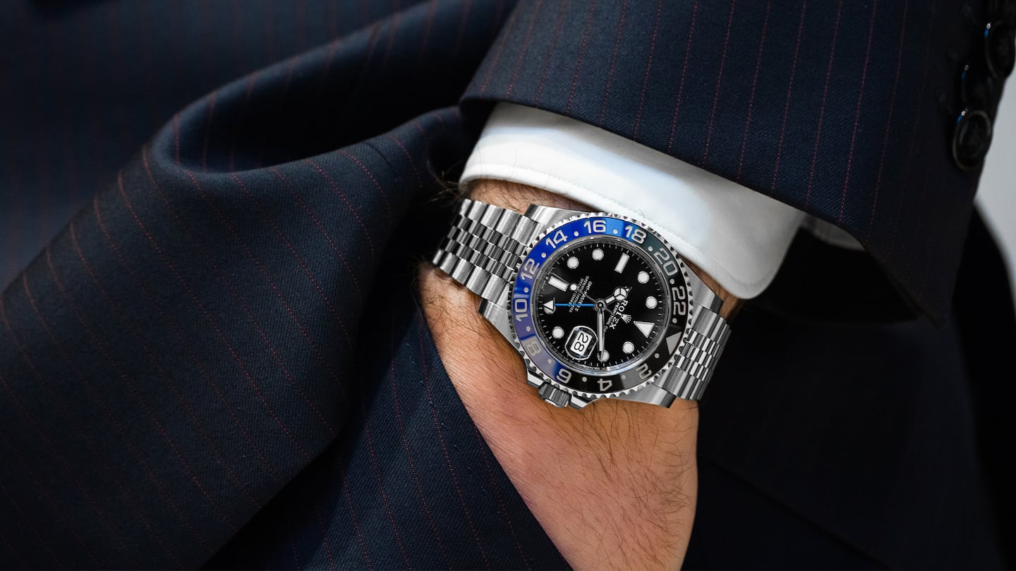 A person wears a silver and blue Rolex watch with a black suit jacket and trousers.