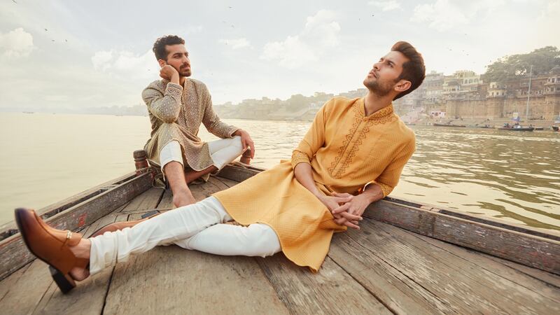 Two models on the edge of a boat wearing traditional Indian garments for men in orange, brown and white.