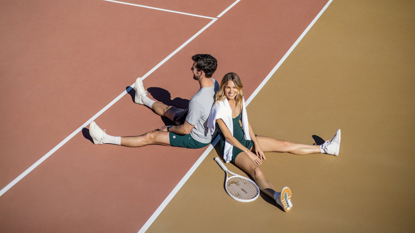 Man and woman sit in middle of tennis court wearing clothing made by Vuori.