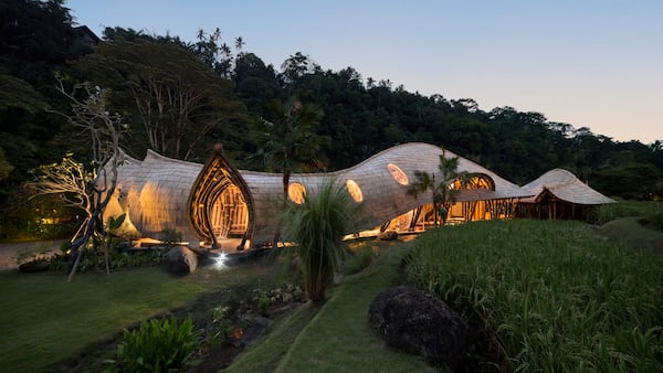 A striking, organic-shaped building sits nestled in lush tropical greenery, its flowing bamboo architecture blending seamlessly with the surrounding landscape. Warm interior lighting glows through oval windows, contrasting beautifully with the cool dusk sky. The structure curves like a natural form—almost wave-like—framed by palm trees, rocks, and manicured grass, with a rice field in the foreground adding to the serene, resort-like atmosphere.