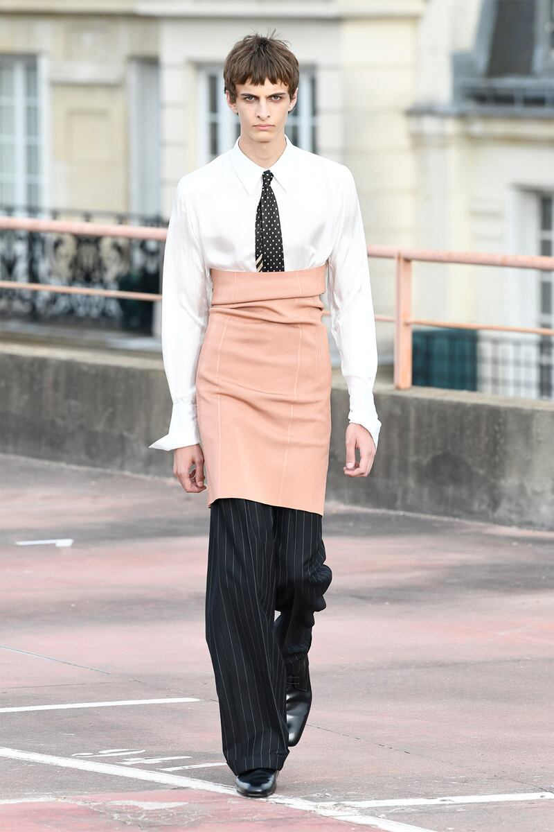 A model wearing a white shirt and tie with pin stripe trousers and formal shoes. Layered over the top is a pale pink pencil skirt.