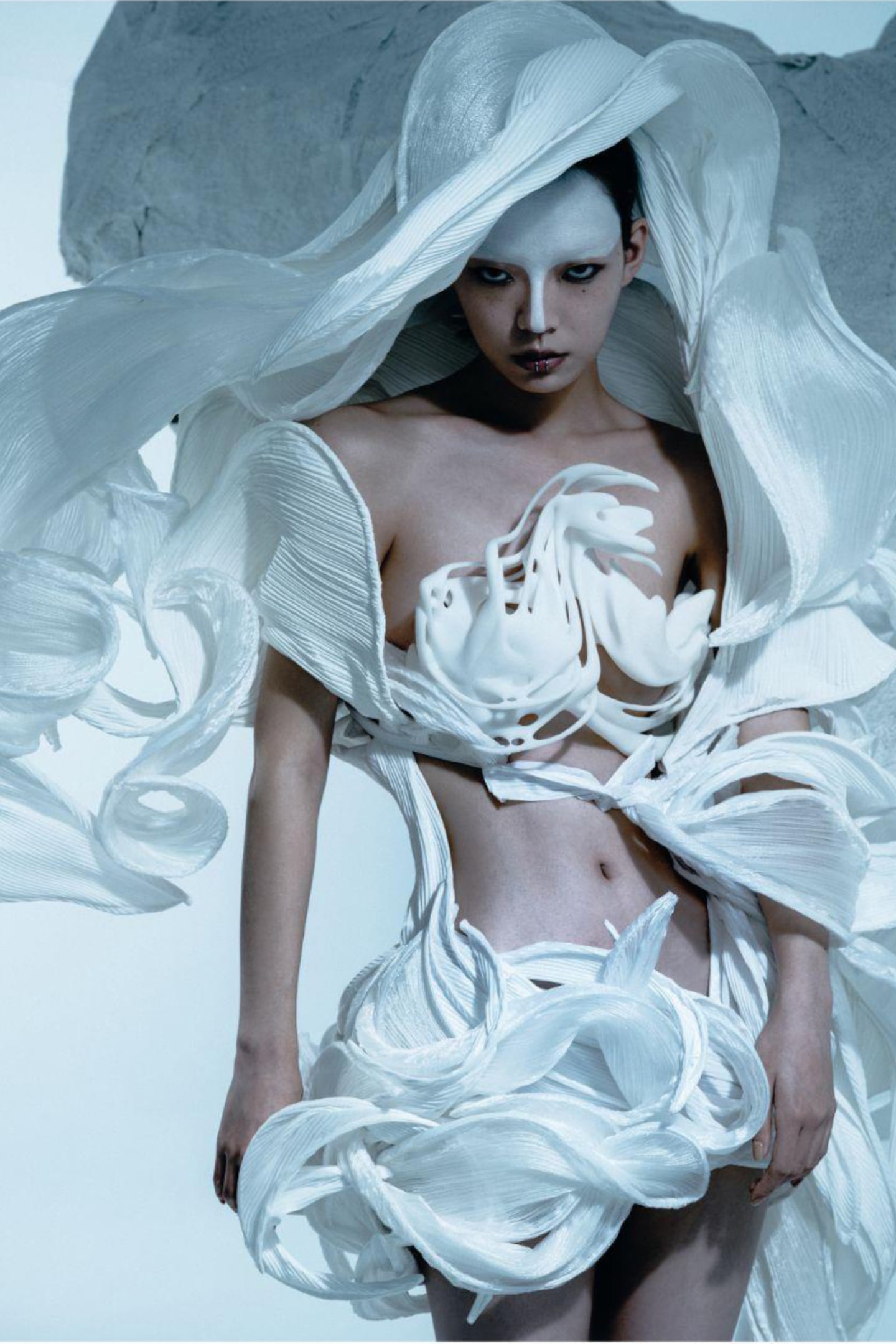A photorealistic digital model stares directly into the camera, dressed in a surreal dress of billowing white fabric.