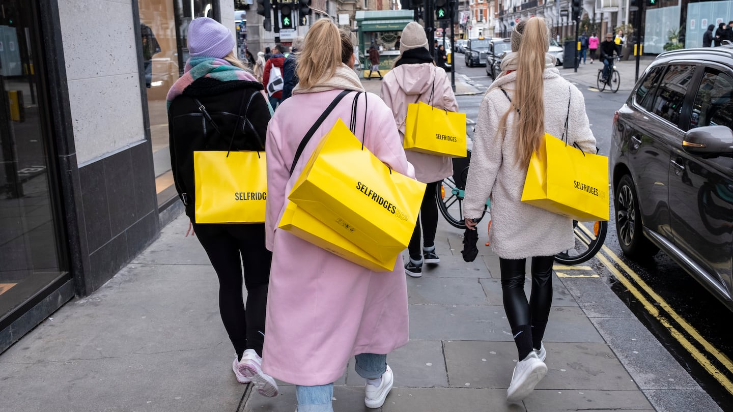 A group of women walk down a busy shopping street carrying large yellow Selfridges bags.