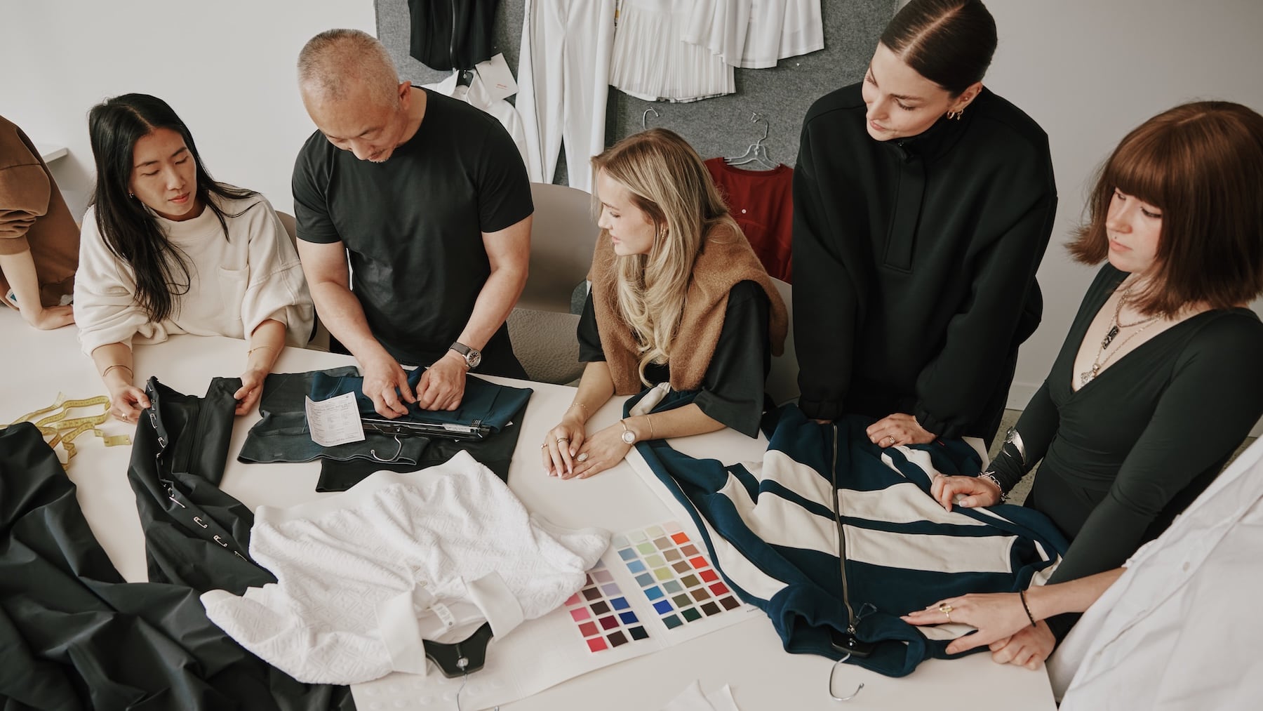 A diverse team of five design professionals gather around a large white table in a brightly lit apparel studio. They are focused on product development, examining various clothing items including a white textured shirt, dark athletic pants, and a blue-and-cream striped jacket. A color palette swatch sheet and a measuring tape rest on the table. In the background, additional garments hang on a design board.