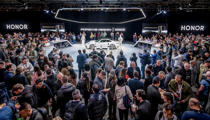 A crowd of people surrounding a central stage where a white Porsche car stands.