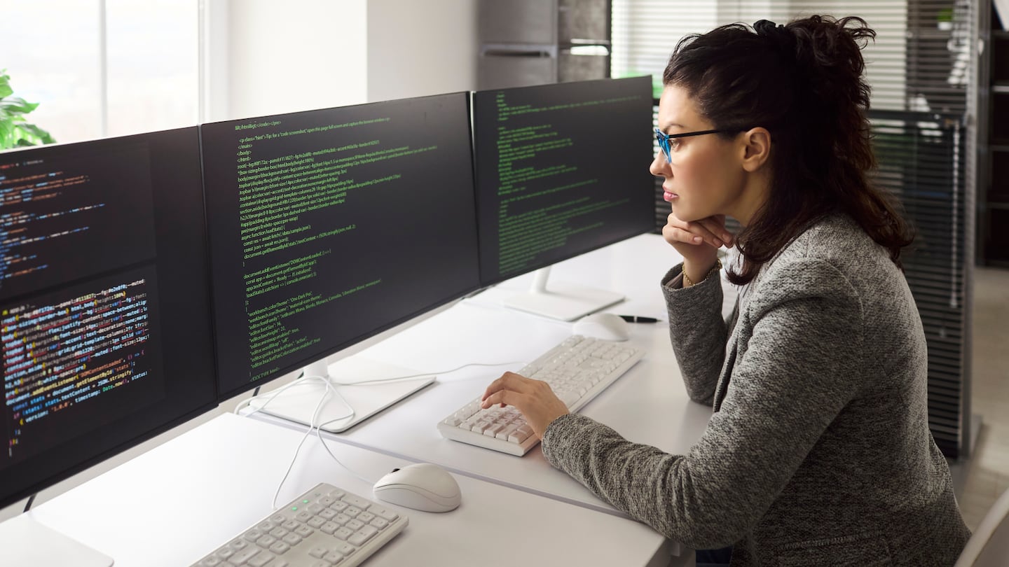 A woman looks at code on three computer monitors.