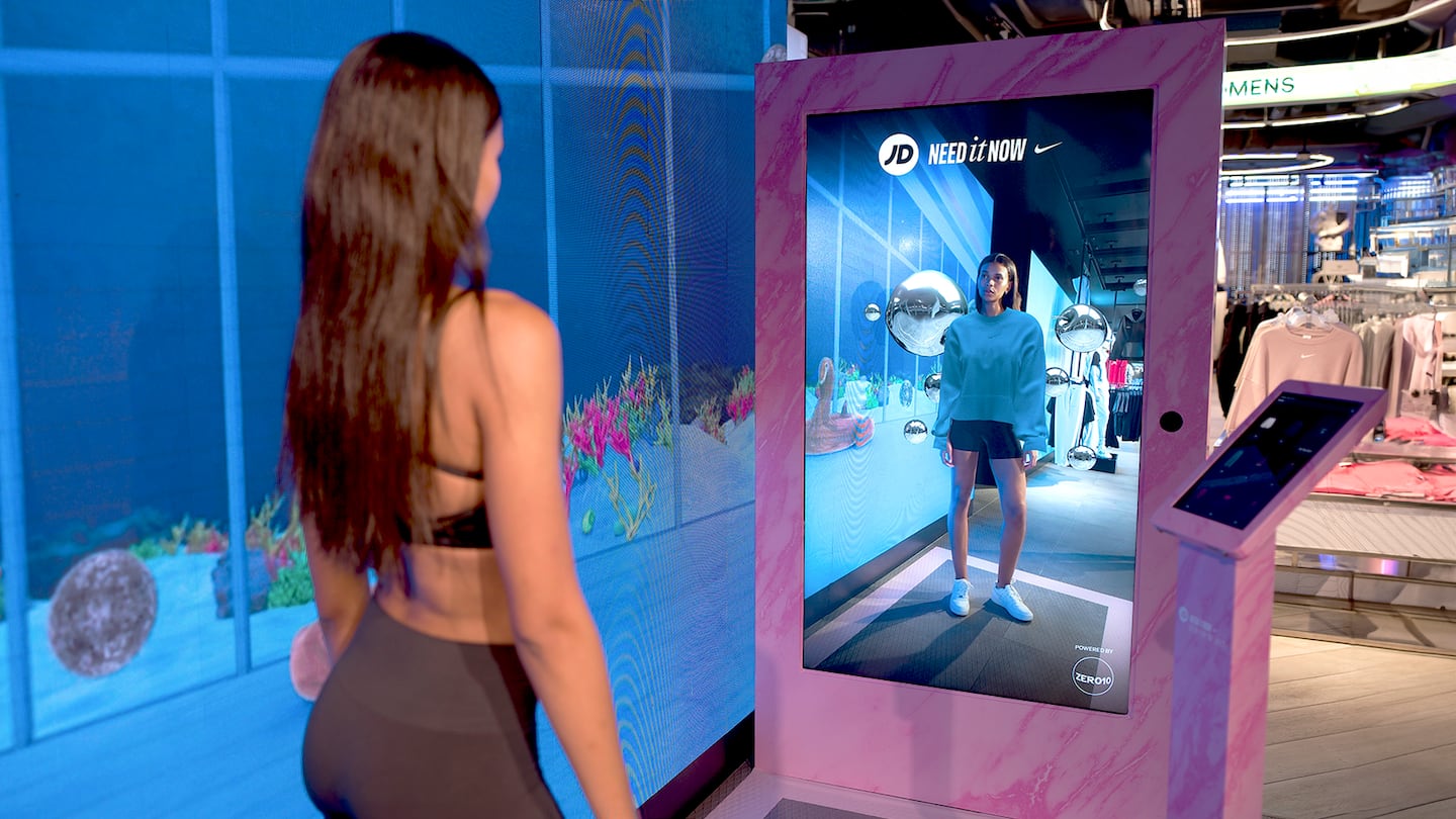 A person stands inside a modern retail store in front of a large digital mirror framed in pink. The mirror displays their reflection wearing virtual clothing, with racks of clothing and other items visible in the background.