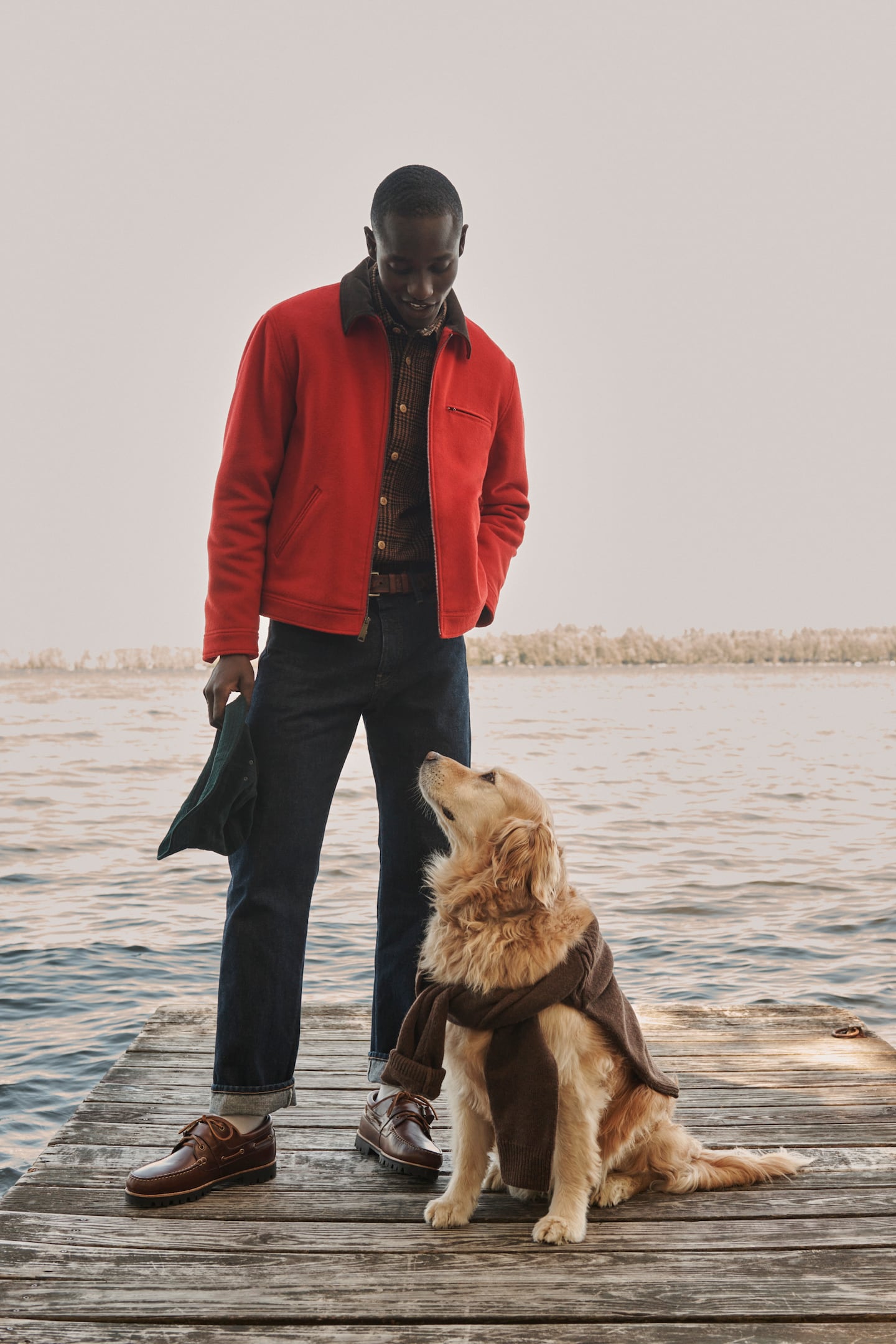 Man in red jacket stands on a dock, looking down to a golden retriever with a sweater tied around its body.