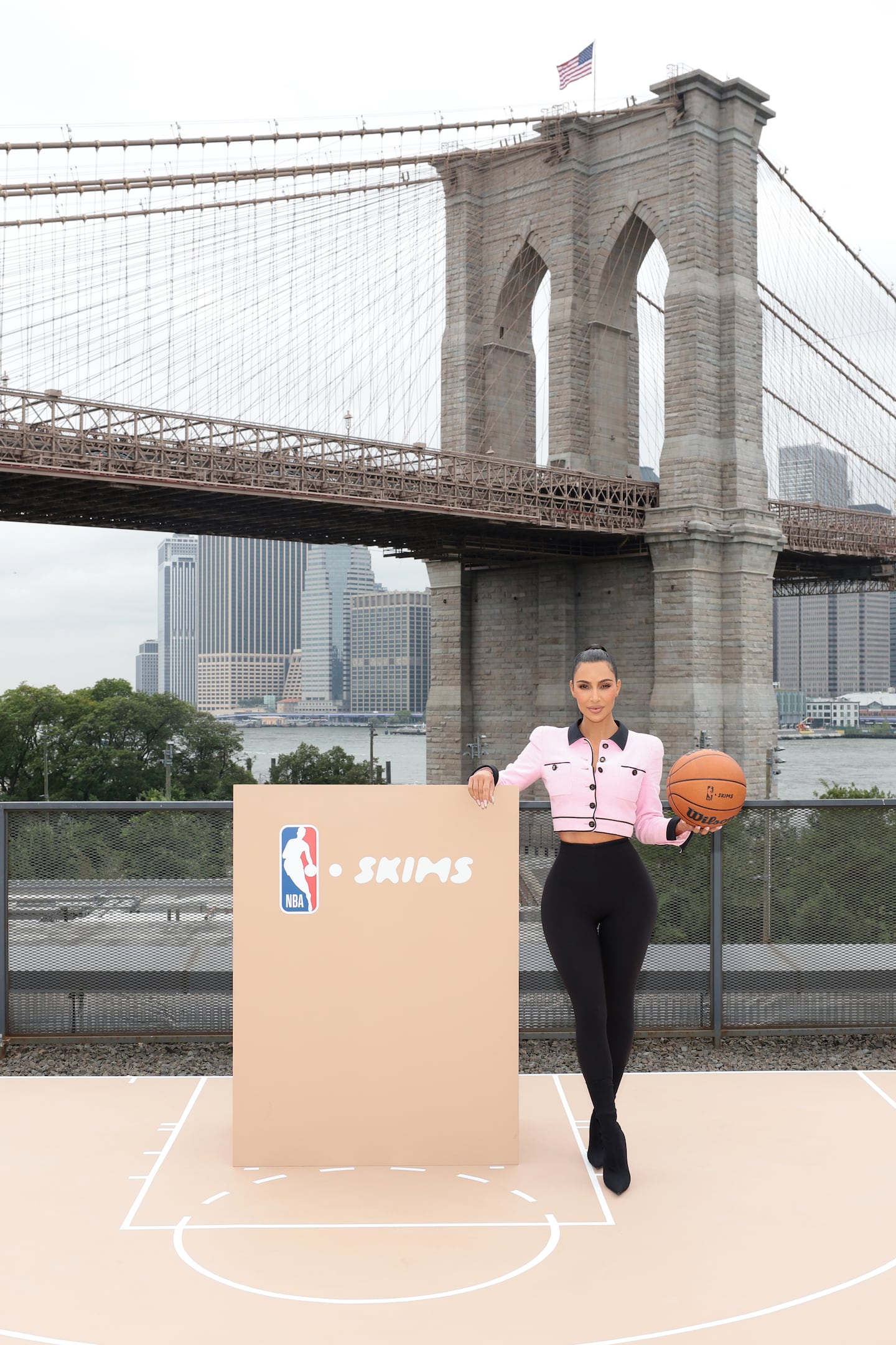 Kim Kardashian stands next to a podium with NBA and Skims signage holding a basketball. The Brooklyn Bridge in New York City is in the background.