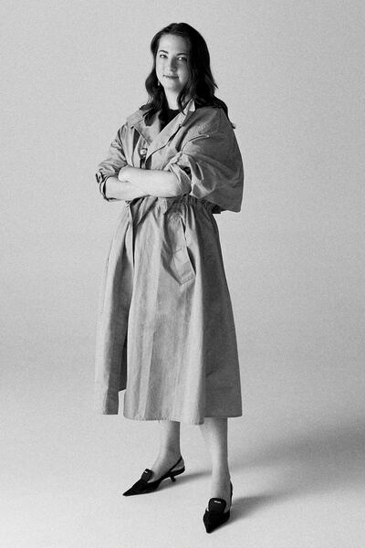 Designer Nensi Dojaka standing in a black and white portrait image, wearing a coat and slingback heels, facing the camera with arms crossed.