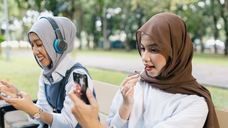 Two Muslim girls sit on a bench, one applies lip gloss and the other listens to music
