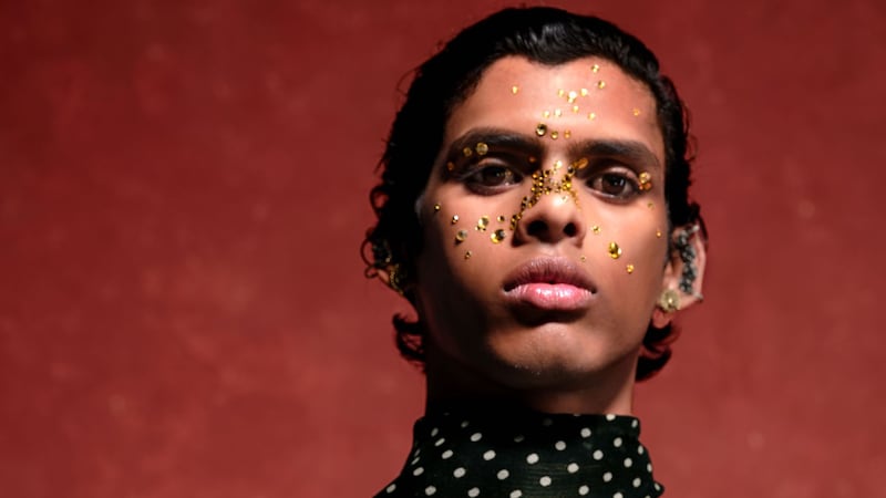A model with glitter on their face stands before a mottled red background.