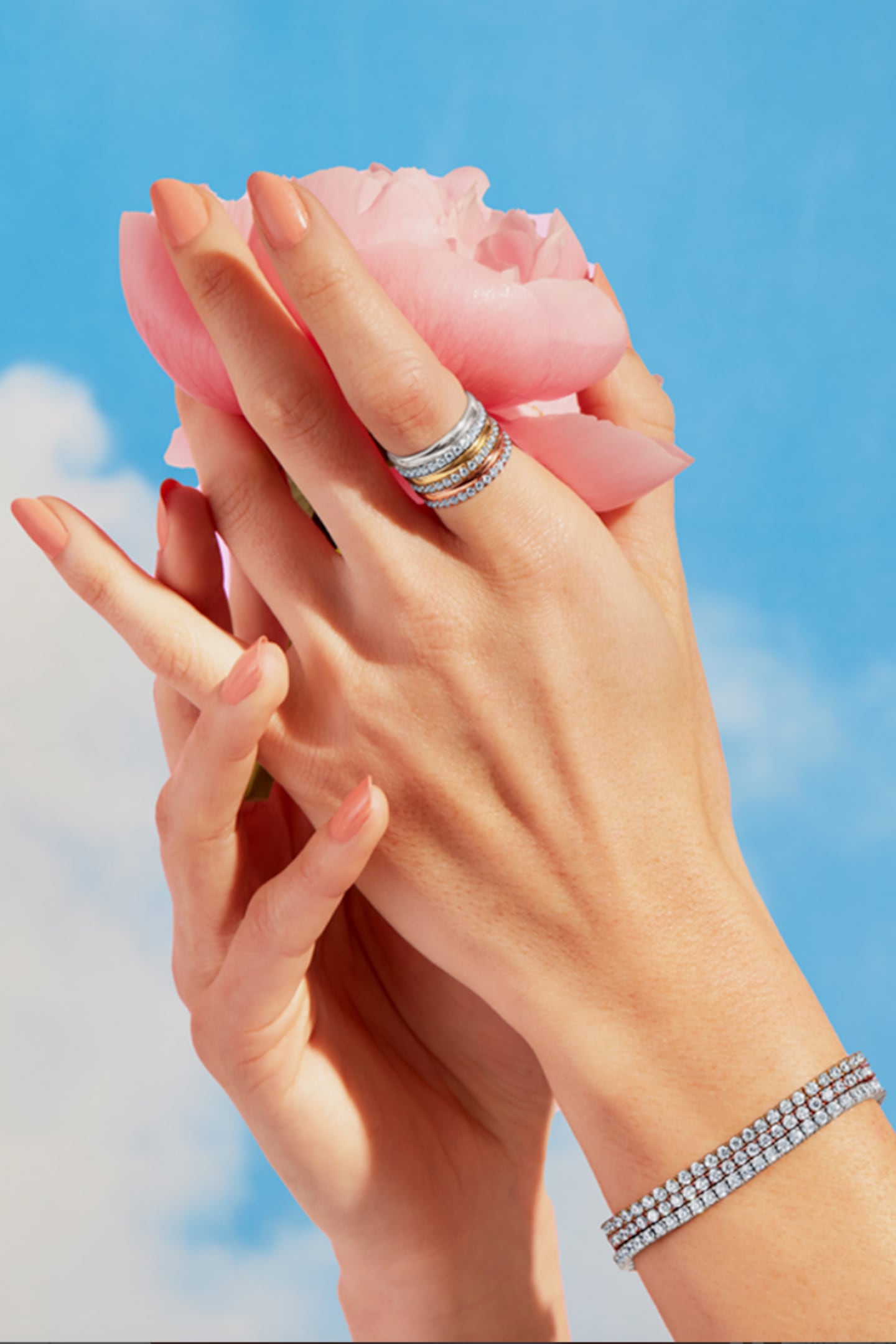 Two hands cup a pink rose against a blue sky. One finger and wrist are bejewelled with diamond-encrusted rings and bracelets.