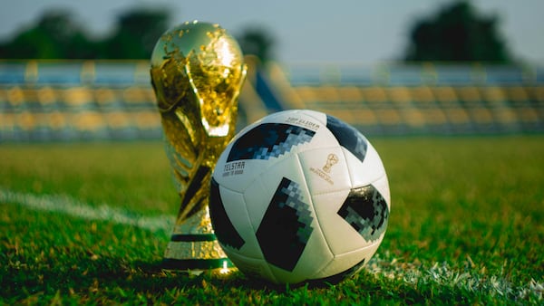 A photo of a soccer ball sitting in front of the World Cup trophy on a grass pitch.