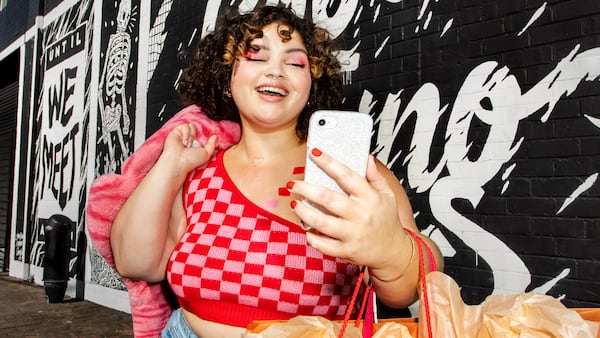 The image shows an individual standing in front of a black and white graffiti wall. They are wearing a red and pink checkered top, blue jeans, and holding a pink coat or sweater over one arm. The individual also has multiple shopping bags, suggesting they have been shopping, and is holding a smartphone in their hand.