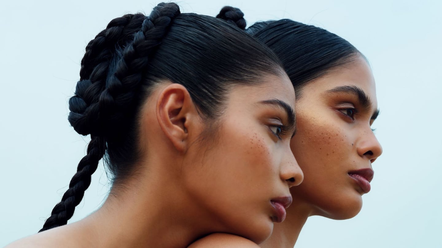 Two women with dark braided hair in profile look into the distance, the woman on the left placing her chin on the other's shoulder.