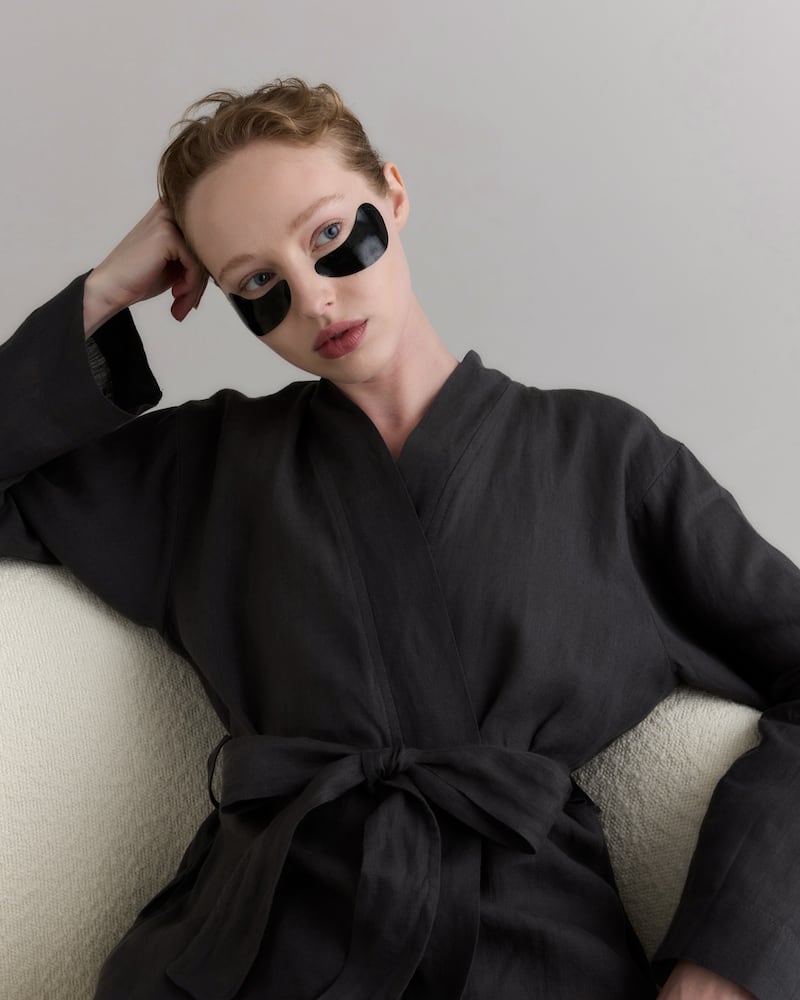 A model wearing a linen robe and 111Skin black undereye patches lounges on a boucle chair