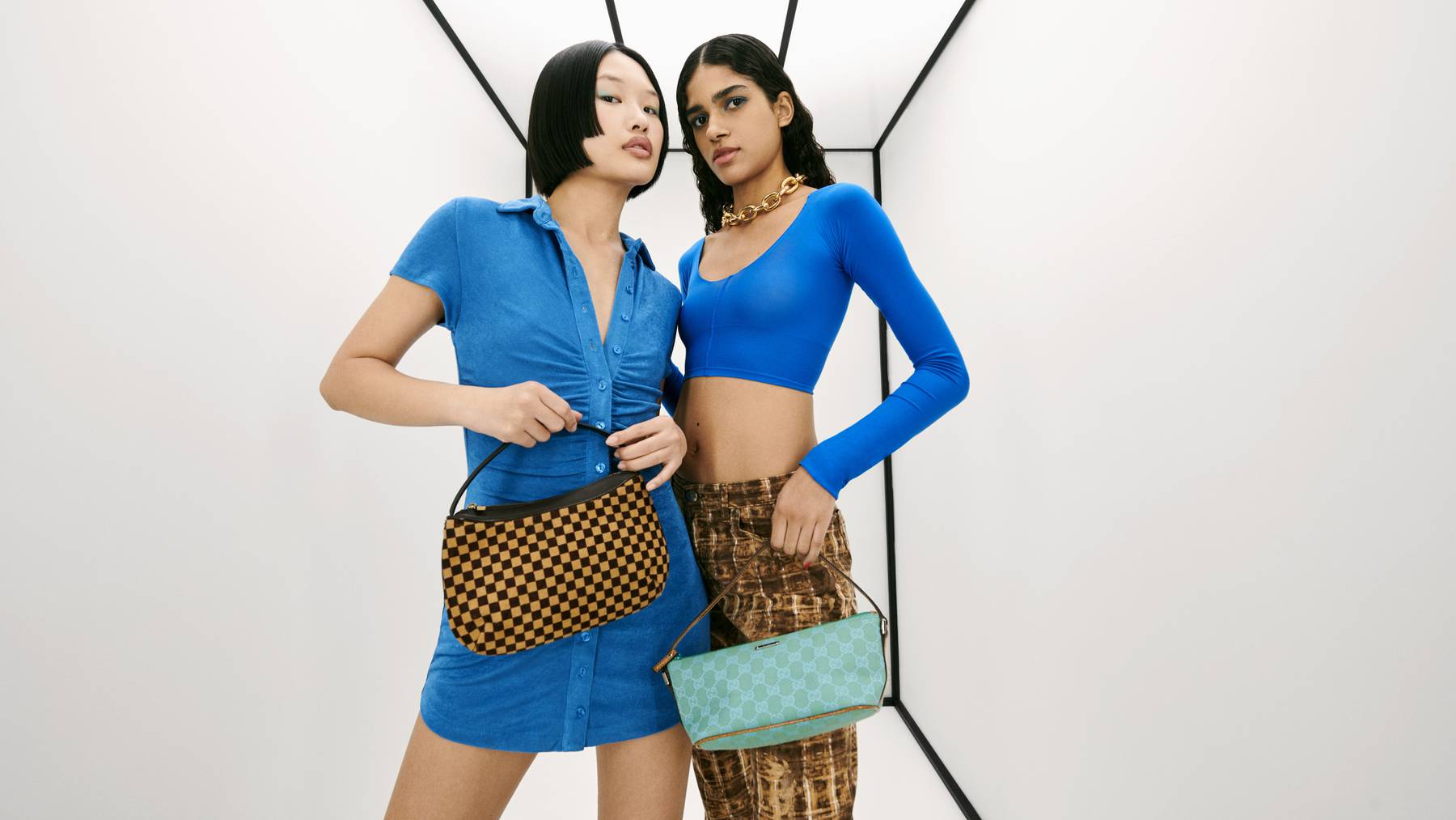 Two models wearing blue standing in a white room holding designer handbags.