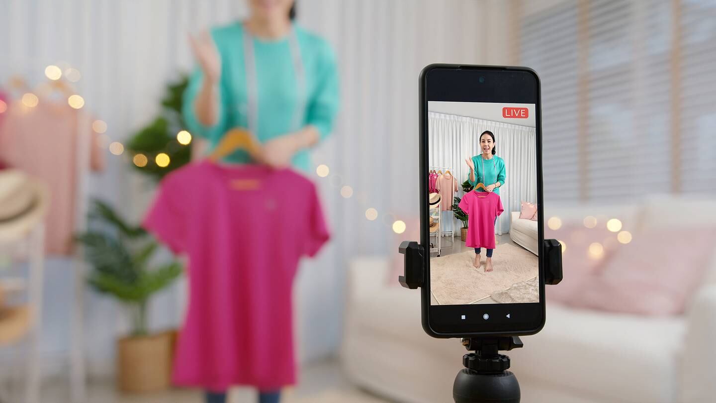 Women livestreaming with a mobile phone on a tripod holding a pink T-shirt.