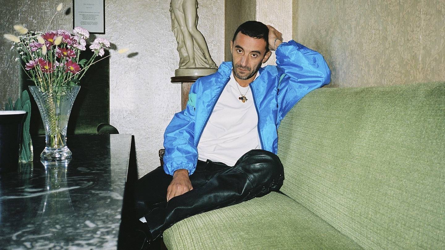 Walter Chiapponi sits on a green sofa wearing a bright blue jacket and black leather trousers.